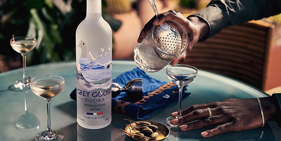 A bottle of Grey Goose Vodka on a glass table next to two short vodka martinis and a third one being poured from a shaker.