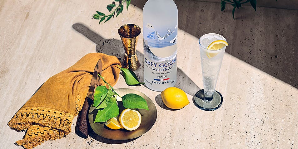 A bottle of Grey Goose Vodka on a counter next to a jigger, a Grey Goose long martini cocktail garnished with lemon, a cut up lemon, leaves for garnish and a knife.