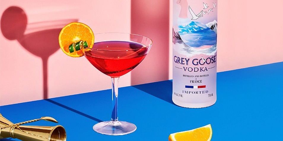 A Grey Goose vodka cosmopolitan cocktail garnished with an orange slice and a lime twist next to a bottle of Grey Goose vodka.