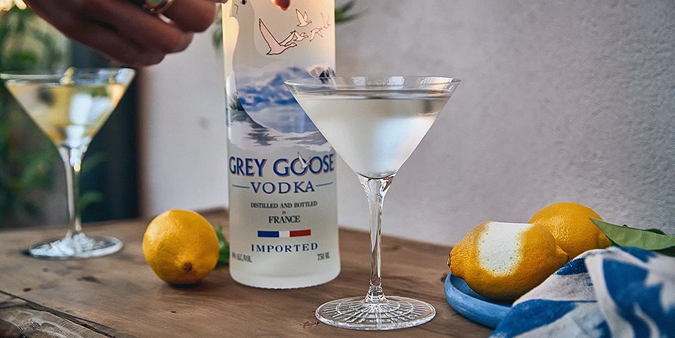 A bottle of Grey Goose Vodka on a wooden table with two classic dry vodka martini cocktails next to a plate of lemons.