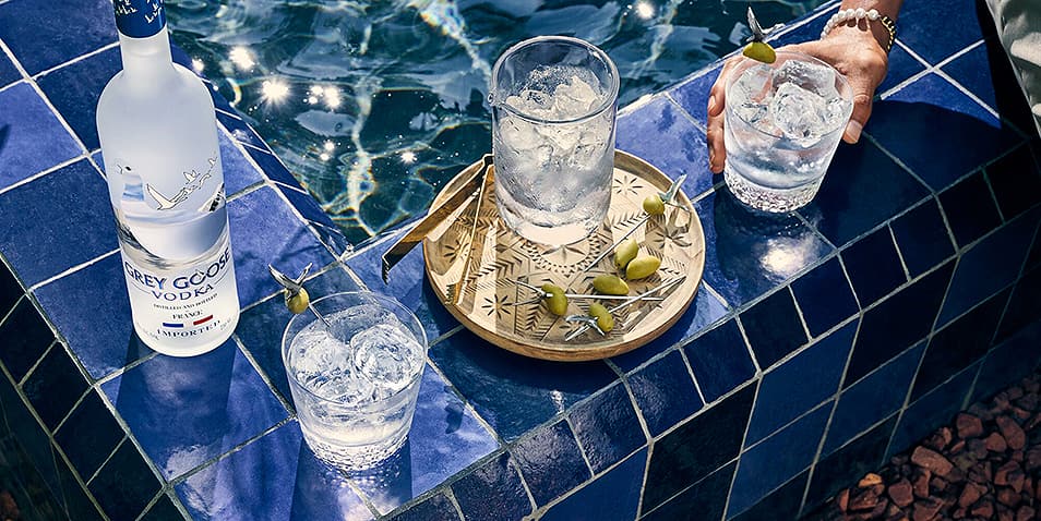 A bottle of Grey Goose Vodka poolside next to three Grey Goose Vodka wet martini cocktails garnished with caper berries on a tray.