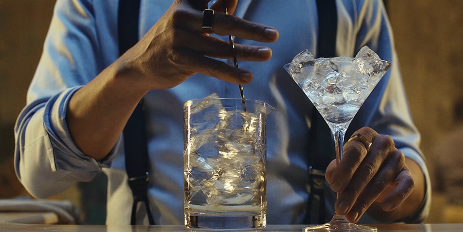 A bartender stirring a cocktail with ice cubes in a rocks glass while also holding a martini glass full of ice.