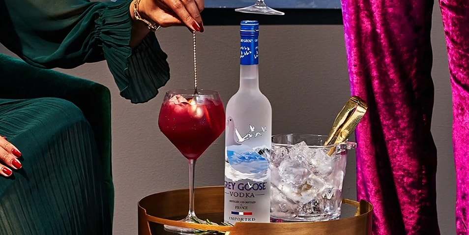 A bottle of Grey Goose Vodka on a table next to a bucket of ice and a glass of Grey Goose holiday punch being stirred with a bar spoon.