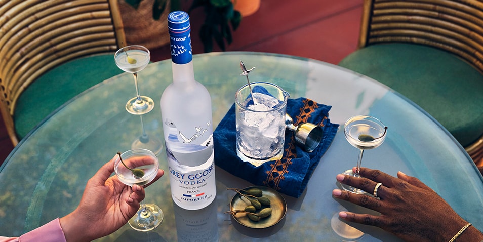 A bottle of Grey Goose Vodka on a table with three vodka martini cocktails garnished with caper berries.
